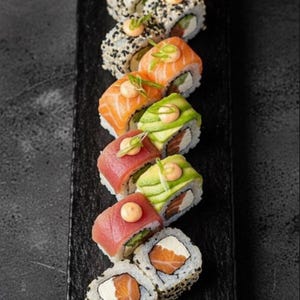 Może przedstawiać: Prostokątna czarna taca z łupka z różnorodnymi rolkami sushi. W zestawie znajdują się rolki z łososiem, tuńczykiem, awokado i czarnym sezamem, każda udekorowana kleksem sosu, szczypiorkiem, imbirem oraz pastą wasabi.