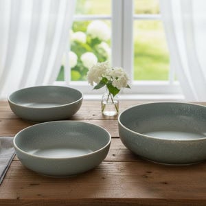 May include: Three gray ceramic bowls of varying sizes are arranged on a wooden table. The bowls have a speckled texture and a slightly irregular shape. A small vase of white flowers sits near a window with sheer curtains.