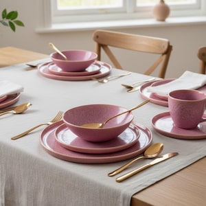 Może przedstawiać: Zestaw stołowy z różowymi ceramicznymi miskami, talerzami i kubkiem. Złote łyżki, widelce i noże są ułożone wokół nakryć. Biała lniana serwetka spoczywa na talerzu.