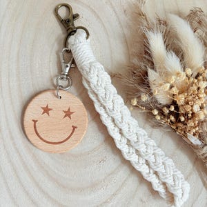 Könnte beinhalten: Ein Holzschlüsselanhänger mit einem Smiley-Gesicht-Design. Der Schlüsselanhänger hat einen runden, hellfarbigen Holzanhänger mit einem lächelnden Gesicht und sternförmigen Augen. Er hat eine weiße geflochtene Handschlaufe und einen Metallverschluss.