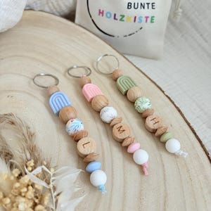 Könnte beinhalten: Drei Holzschlüsselanhänger mit verschiedenen Perlen und Anhängern. Jeder Schlüsselanhänger hat ein anderes Farbschema, darunter Blau, Rosa und Grün. Ein Schlüsselanhänger hat den Buchstaben "L", ein anderer "M" und der letzte "H". Im Hintergrund befindet sich ein kleiner Beutel mit dem Text "DIE BUNTE HOLZKISTE".
