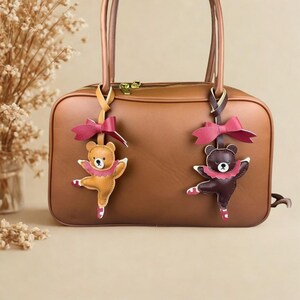 Può includere: Una borsa in pelle marrone con due ciondoli a forma di orso. I ciondoli sono orsi danzanti, uno marrone chiaro e uno marrone scuro, con dettagli rosa e fiocchi rossi. La borsa ha una chiusura a zip.