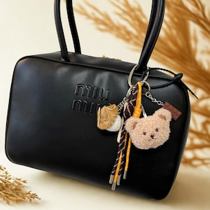 Puede incluir: Bolso rectangular negro con logo en relieve. El bolso tiene dos asas y un llavero decorativo con un guante de béisbol, una pelota de béisbol, un oso de peluche y un pequeño bloque marrón.