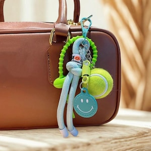 Könnte beinhalten: Ein farbenfroher Schlüsselanhänger mit einem Tennisball, einem grünen Smiley-Anhänger und einem hellblauen Seildesign. Der Schlüsselanhänger ist an einer braunen Handtasche mit goldenem Griff befestigt.
