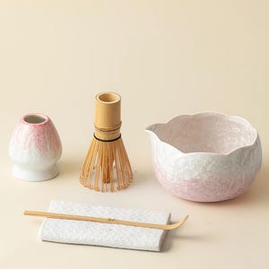 Op de afbeelding: Een matcha-theeset met een roze en witte keramische kom, een bamboe klopper, een kleine keramische container, een bamboe lepel en een witte keramische rust. Ontworpen voor het bereiden van traditionele Japanse matcha-thee.