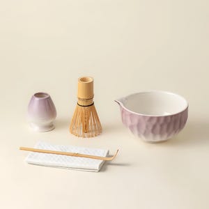 Puede incluir: Un juego de té matcha con un cuenco de cerámica morado y blanco, un batidor de bambú, un pequeño recipiente de cerámica morado, una cuchara de bambú y un paño blanco. El cuenco tiene un exterior texturizado.