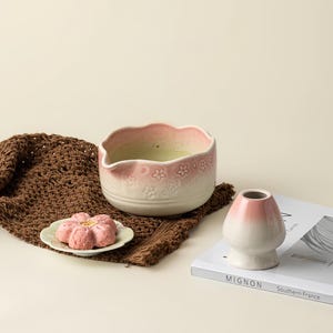 May include: A ceramic matcha bowl with a pink rim and floral detailing, filled with green matcha. A small pink flower-shaped pastry sits on a white plate. A white and pink chasen holder and a book are also present.