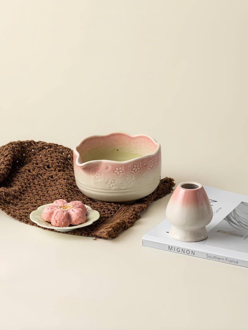 May include: A ceramic matcha bowl with a pink rim and floral detailing, filled with green matcha. A small pink flower-shaped pastry sits on a white plate. A white and pink chasen holder and a book are also present.