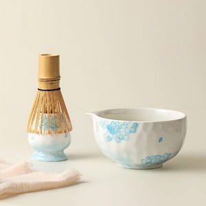 Può includere: Un set da tè matcha con una frusta di bambù e una ciotola di ceramica bianca con beccuccio. La ciotola e il supporto della frusta sono decorati con motivi floreali blu. La frusta è in bambù.