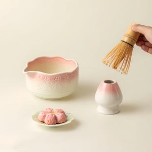 May include: A matcha tea set comprising a white and pink ceramic bowl with a floral pattern, a bamboo whisk, a white and pink ceramic whisk holder, and a pink flower-shaped pastry on a small plate. The items are set against a light background.