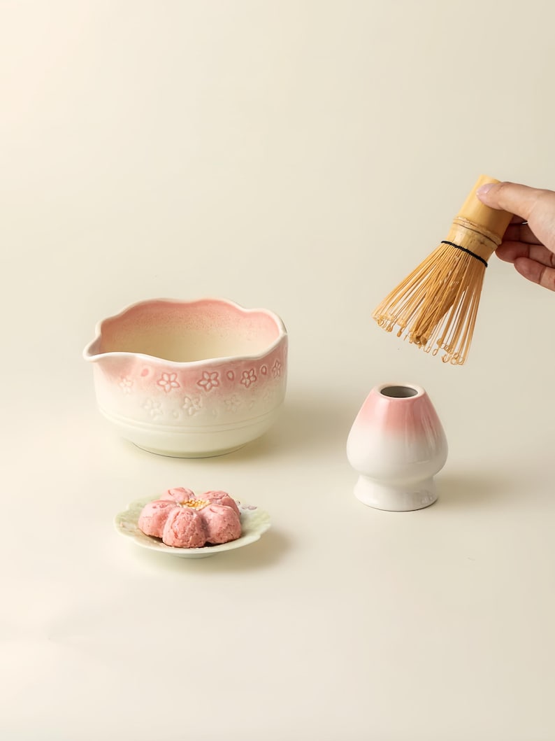 May include: A matcha tea set comprising a white and pink ceramic bowl with a floral pattern, a bamboo whisk, a white and pink ceramic whisk holder, and a pink flower-shaped pastry on a small plate. The items are set against a light background.