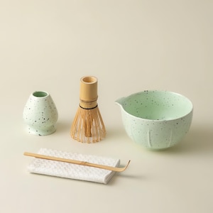 May include: A matcha tea set comprising a pale green ceramic bowl and vase with a speckled pattern, a bamboo whisk, and a bamboo scoop on a white cloth. The set is designed for preparing matcha tea.