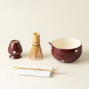 May include: A matcha tea set featuring a maroon ceramic bowl, a bamboo whisk, a bamboo scoop, a ceramic whisk holder, and a white cloth. The bowl and holder are decorated with white flower accents.