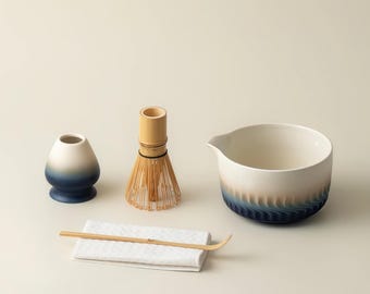 Puede incluir: Un juego de té matcha con un cuenco de cerámica blanco y azul, un batidor de bambú, un pequeño recipiente de cerámica, una cuchara de bambú y un paño blanco. El cuenco y el recipiente tienen un degradado azul.