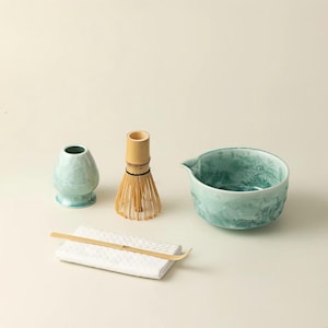 Puede incluir: Un juego de té matcha con un cuenco de cerámica azul claro, un batidor, una cuchara, un jarrón pequeño y un paño blanco. El cuenco tiene una superficie texturizada y un pico. El batidor es de bambú. El juego es para té matcha.