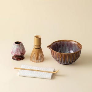 May include: A matcha tea set featuring a ceramic bowl with a spout, a bamboo whisk, a whisk holder, a scoop, and a ceramic whisk stand. The bowl is brown and purple, and the other items are natural wood and white.