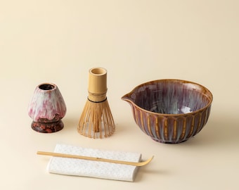 May include: A matcha tea set featuring a ceramic bowl with a spout, a bamboo whisk, a whisk holder, a scoop, and a ceramic whisk stand. The bowl is brown and purple, and the other items are natural wood and white.