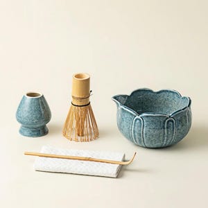 May include: A matcha tea set featuring a blue ceramic bowl, a whisk, a scoop, and a small ceramic container. The set includes a bamboo whisk and scoop, and a textured white stone rest.