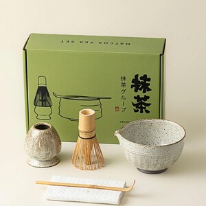 May include: A matcha tea set with a green box that reads "Matcha Tea Set" in English and Japanese. The set includes a ceramic bowl, whisk, scoop, whisk holder, and a small ceramic container. The bowl is speckled with a dark rim.