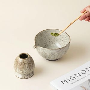 May include: A speckled ceramic matcha bowl with a spout, a small ceramic matcha whisk holder, and a bamboo matcha spoon holding green matcha powder. A book with the word "MIGNON" is in the background. The items are on a light beige surface.