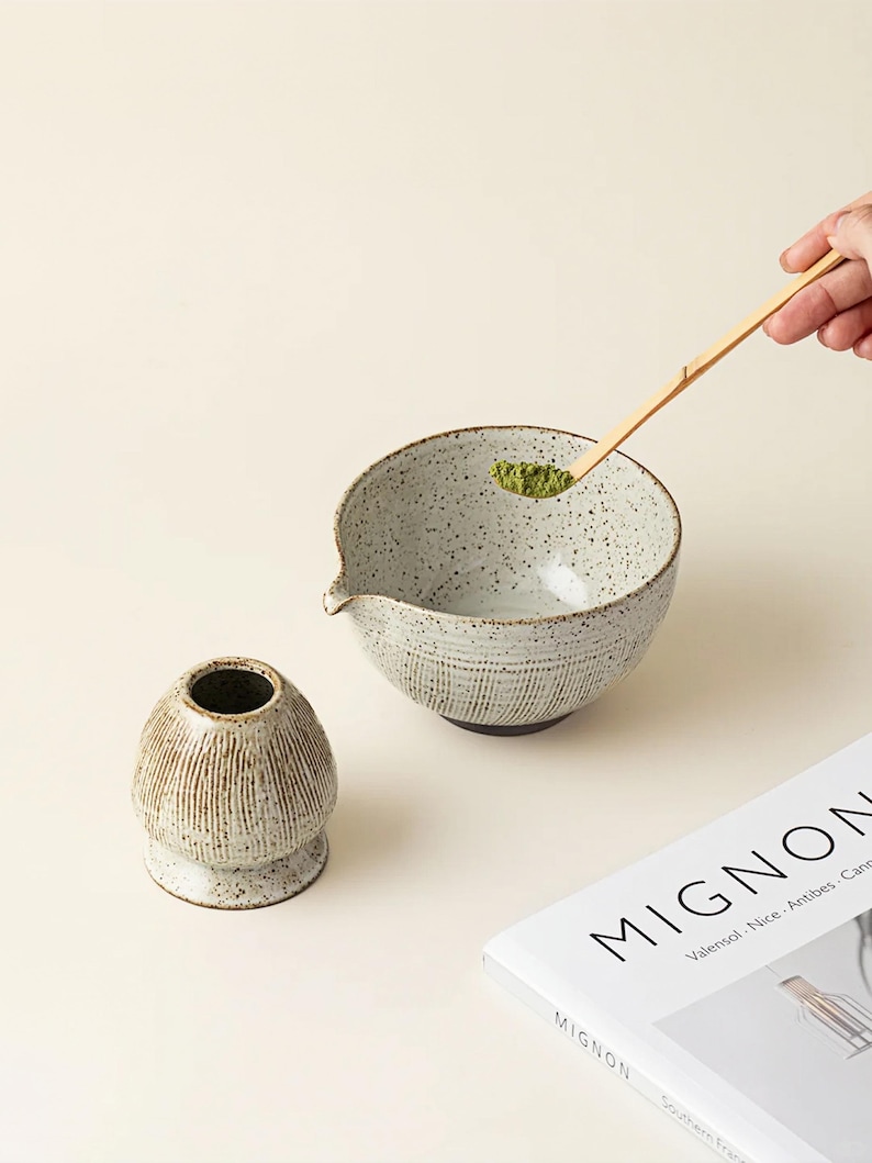 May include: A speckled ceramic matcha bowl with a spout, a small ceramic matcha whisk holder, and a bamboo matcha spoon holding green matcha powder. A book with the word "MIGNON" is in the background. The items are on a light beige surface.