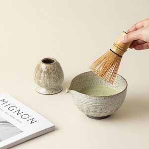 May include: A matcha tea set with a ceramic bowl, whisk, and tea container. The bowl contains green tea and is being whisked with a bamboo whisk. A magazine with the word "MIGNON" is also in the image.