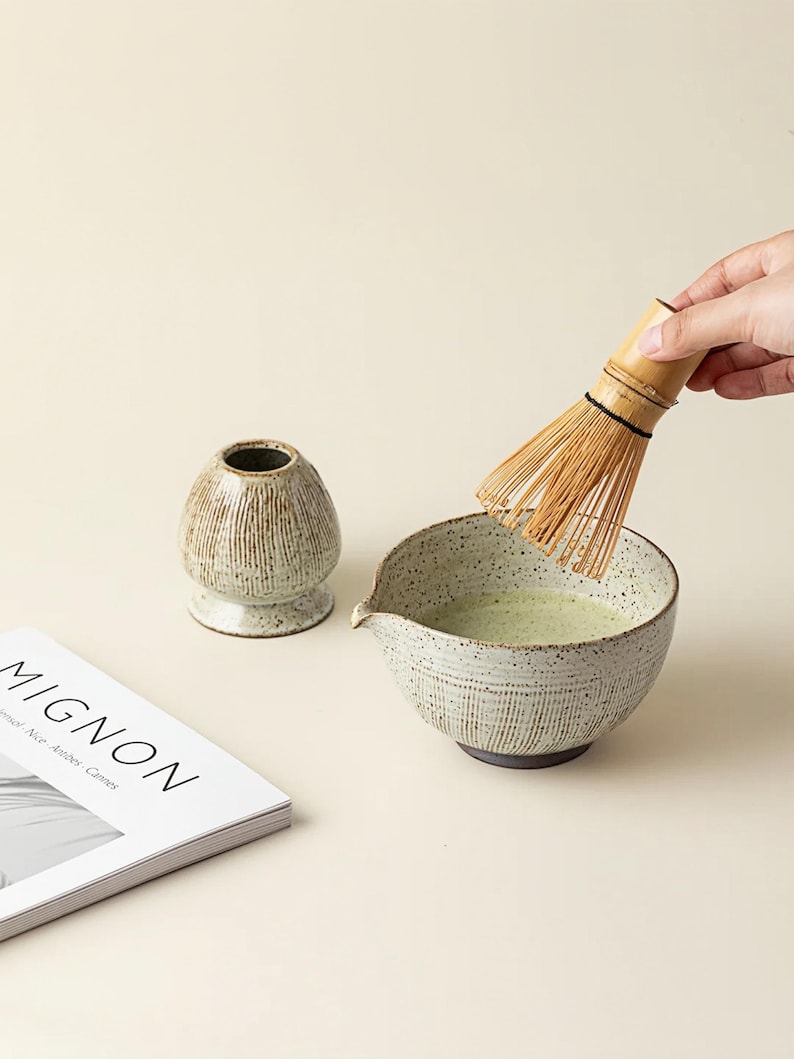 May include: A matcha tea set with a ceramic bowl, whisk, and tea container. The bowl contains green tea and is being whisked with a bamboo whisk. A magazine with the word "MIGNON" is also in the image.