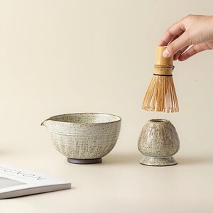 May include: A ceramic matcha bowl with a spout, a whisk, and a whisk holder. The bowl and holder are speckled with a textured, vertical line design. The whisk is made of bamboo. The items are on a neutral background.