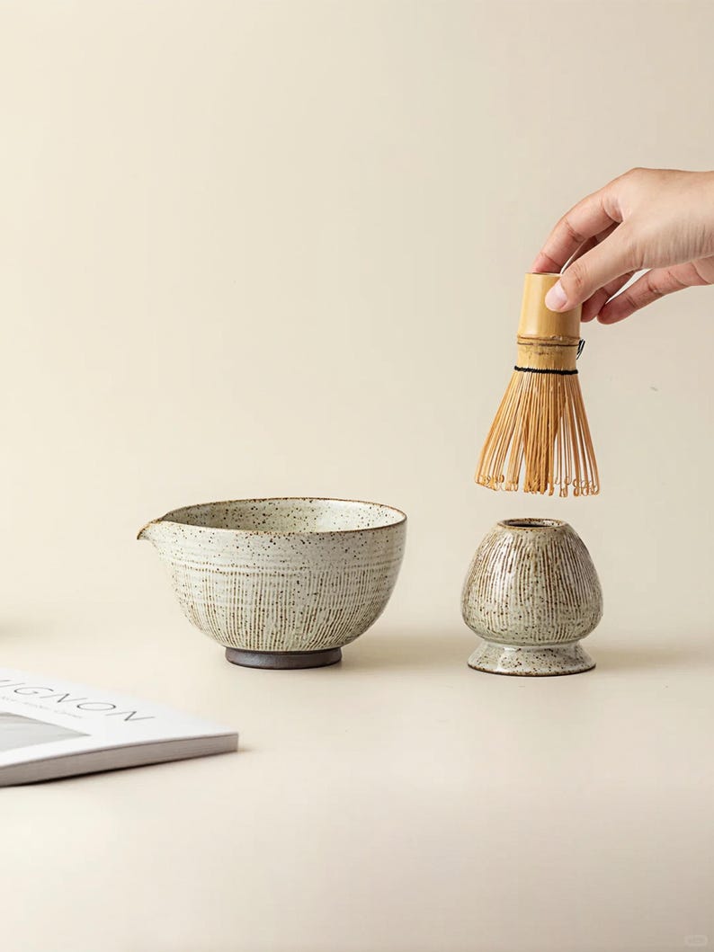 May include: A ceramic matcha bowl with a spout, a whisk, and a whisk holder. The bowl and holder are speckled with a textured, vertical line design. The whisk is made of bamboo. The items are on a neutral background.
