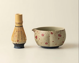 Op de afbeelding: Een bamboe matcha klopper en een keramische chawan theekom. De kom is beige met rode bloempatronen en een blauwe basis. De klopper heeft een beige handvat en een donkerblauwe basis.