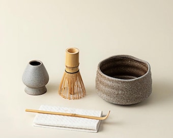 Può includere: Un set da tè matcha con una ciotola in ceramica marrone screziata, un frullino di bambù, un cucchiaio e un piccolo contenitore in ceramica. Il frullino è fatto di bambù colorato chiaro. Il set è disposto su un panno bianco.