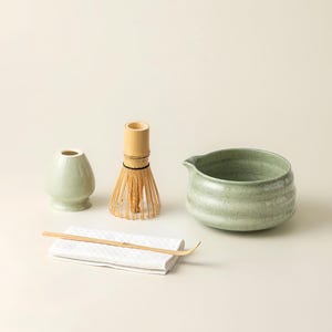 Może przedstawiać: Zestaw do herbaty matcha składający się z jasnoniebieskiej ceramicznej miski, bambusowej miotełki, małego ceramicznego pojemnika, bambusowej łyżki i białej ściereczki. Zestaw do przygotowywania herbaty matcha.