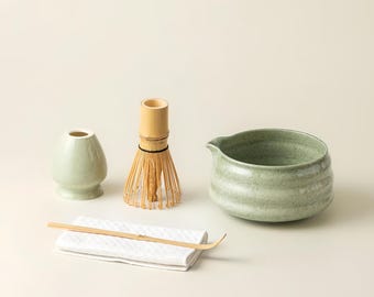 May include: A matcha tea set featuring a light green ceramic bowl, a bamboo whisk, a small ceramic container, a bamboo scoop, and a white cloth. The set is designed for preparing matcha tea.