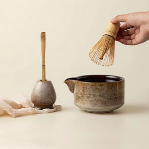 Puede incluir: Un juego de té matcha con un cuenco de cerámica con boquilla, un batidor de bambú y un soporte para batidor con una cuchara de bambú. El cuenco y el soporte tienen un tono moteado y terroso. El batidor se sostiene sobre el cuenco.