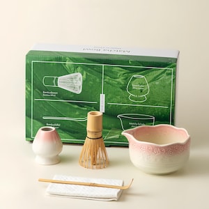 May include: A matcha tea set including a bamboo whisk, a ceramic bowl with a floral pattern, a bamboo scoop, a whisk holder, and a white cloth. The box has a green background with illustrations and the words "Matcha Bowl".