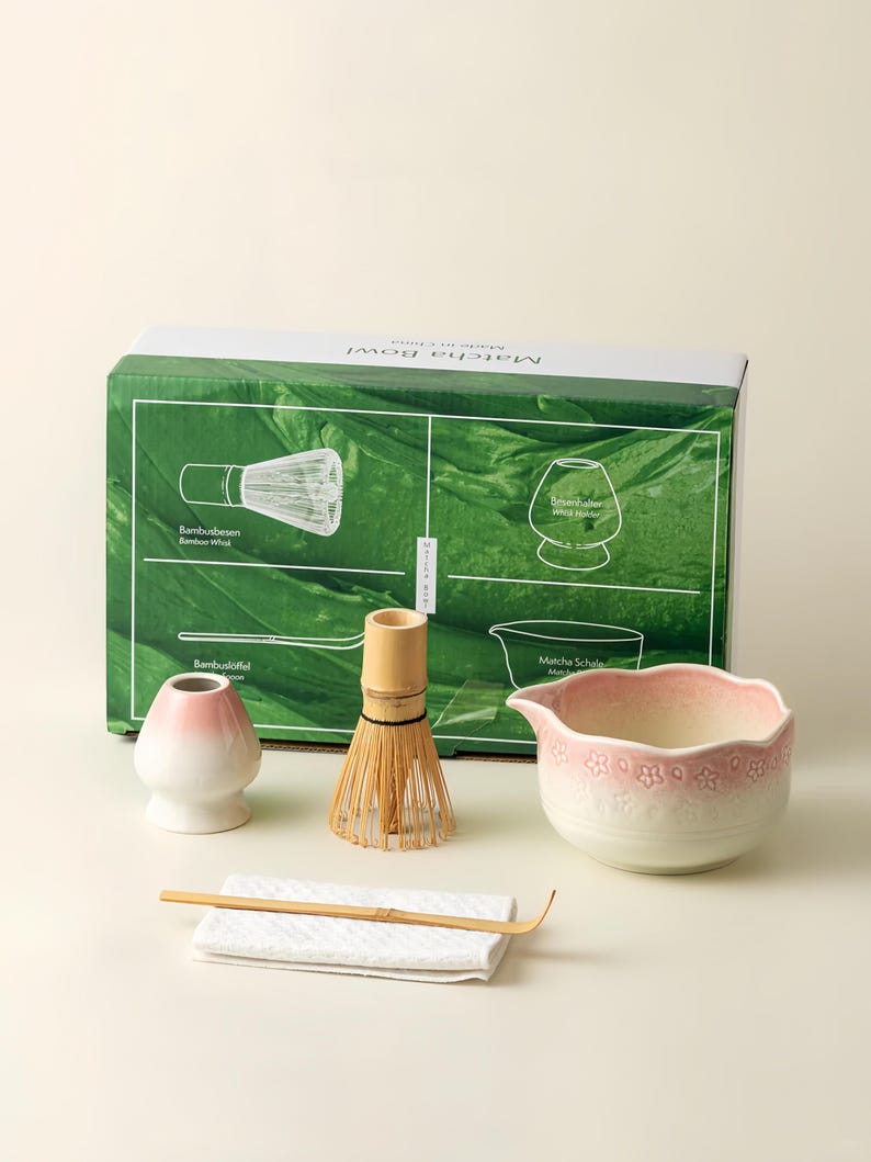 May include: A matcha tea set including a bamboo whisk, a ceramic bowl with a floral pattern, a bamboo scoop, a whisk holder, and a white cloth. The box has a green background with illustrations and the words "Matcha Bowl".
