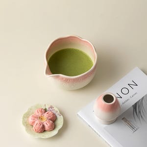 May include: A matcha bowl with a pink floral pattern, containing green matcha tea, is next to a small pink and white vase. A flower-shaped pastry sits on a small plate. A magazine with the word "NON" is also in the image.