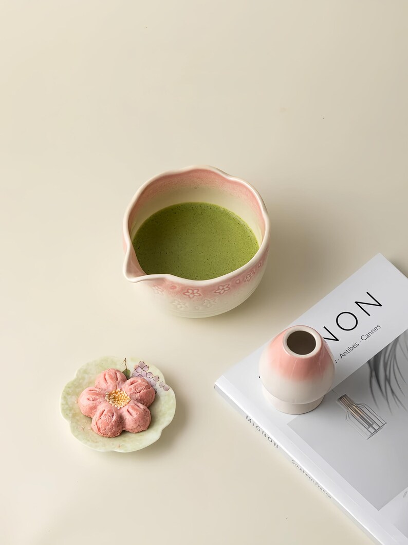 May include: A matcha bowl with a pink floral pattern, containing green matcha tea, is next to a small pink and white vase. A flower-shaped pastry sits on a small plate. A magazine with the word "NON" is also in the image.