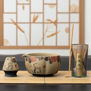 Pode incluir: Um conjunto de chá de cerâmica bege com um design de gato e floral. O conjunto inclui uma tigela, um recipiente pequeno, um batedor de bambu e um suporte para batedor. A tigela tem um bico e é decorada com flores vermelhas e pretas.