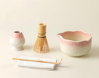 May include: A matcha tea set featuring a pink and white ceramic bowl, a bamboo whisk, a bamboo scoop, a white ceramic tea bowl, and a white cloth. The bowl has a floral design around the rim. The whisk is made of thin bamboo tines.