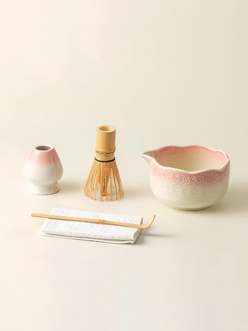 May include: A matcha tea set comprising a pink and white ceramic bowl, a bamboo whisk, a bamboo scoop, a white ceramic tea bowl, and a white cloth. The bowl has a floral design around the rim. The whisk is made of thin bamboo tines.