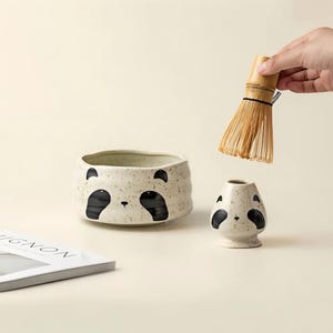 May include: A ceramic matcha bowl and whisk holder set with a panda bear design. The off-white bowls have black panda face details. A bamboo whisk is held above the set. A magazine is in the background.