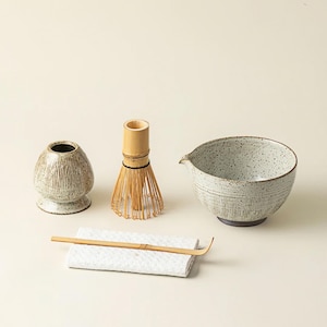 May include: A matcha tea set featuring a ceramic bowl, a bamboo whisk, a whisk holder, a bamboo scoop, and a small ceramic container. The bowl and container have a textured, speckled finish. The whisk is made of bamboo.