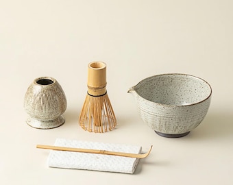 Puede incluir: Un juego de té matcha con un cuenco de cerámica, un batidor de bambú, un soporte para batidor, una cuchara de bambú y un pequeño recipiente de cerámica. El cuenco y el recipiente tienen un acabado texturizado y moteado. El batidor es de bambú.