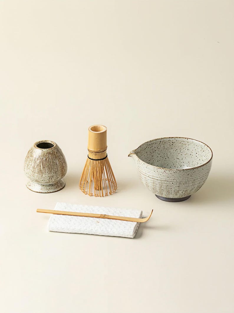 May include: A matcha tea set featuring a ceramic bowl, a bamboo whisk, a whisk holder, a bamboo scoop, and a small ceramic container. The bowl and container have a textured, speckled finish. The whisk is made of bamboo.
