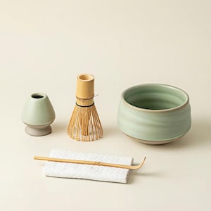 Puede incluir: Un juego de té matcha con un cuenco de cerámica verde claro, un batidor de bambú, un pequeño soporte de cerámica y una cuchara de bambú sobre un paño blanco. El juego es para preparar té matcha japonés tradicional.