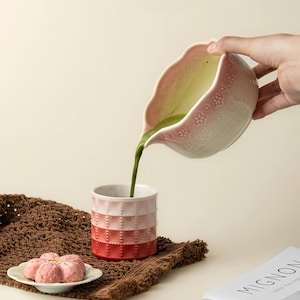 May include: A pink and white ceramic pitcher pours green liquid into a matching ombre cup. The cup transitions from white to red. A small plate holds a pink flower-shaped pastry. A brown woven mat and a book titled "Mignon" are also present.