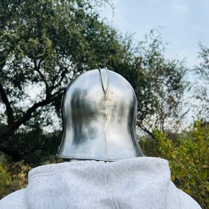 May include: A polished, silver-colored metal helmet, likely for historical reenactment or display. The helmet has a rounded top with a central ridge and a flared base. The background features a person wearing a gray hoodie, with trees and a cloudy sky.