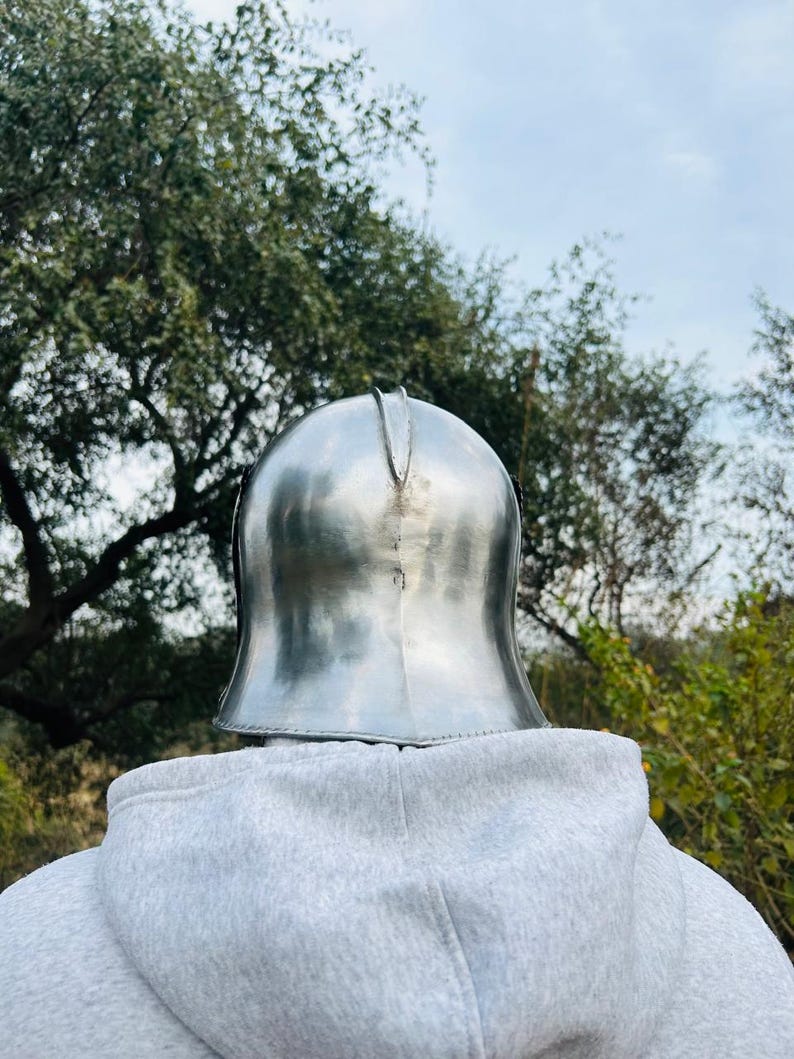 May include: A polished, silver-colored metal helmet, likely for historical reenactment or display. The helmet has a rounded top with a central ridge and a flared base. The background features a person wearing a gray hoodie, with trees and a cloudy sky.