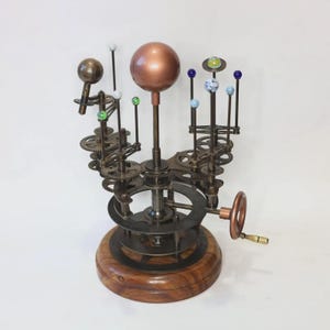 May include: A detailed, steampunk-style model of the solar system. The model features a copper-colored sun, planets represented by colorful glass spheres, and intricate gear mechanisms. The base is made of wood, and a hand crank is visible.